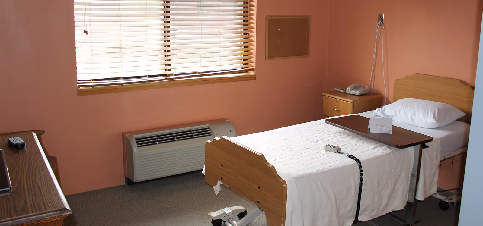 A simple senior care facility bedroom with a single bed covered in a white blanket, a pillow, and an overbed table. The room has peach-colored walls, a window with blinds, a wooden nightstand with a telephone, and a heating or air conditioning unit below the window.