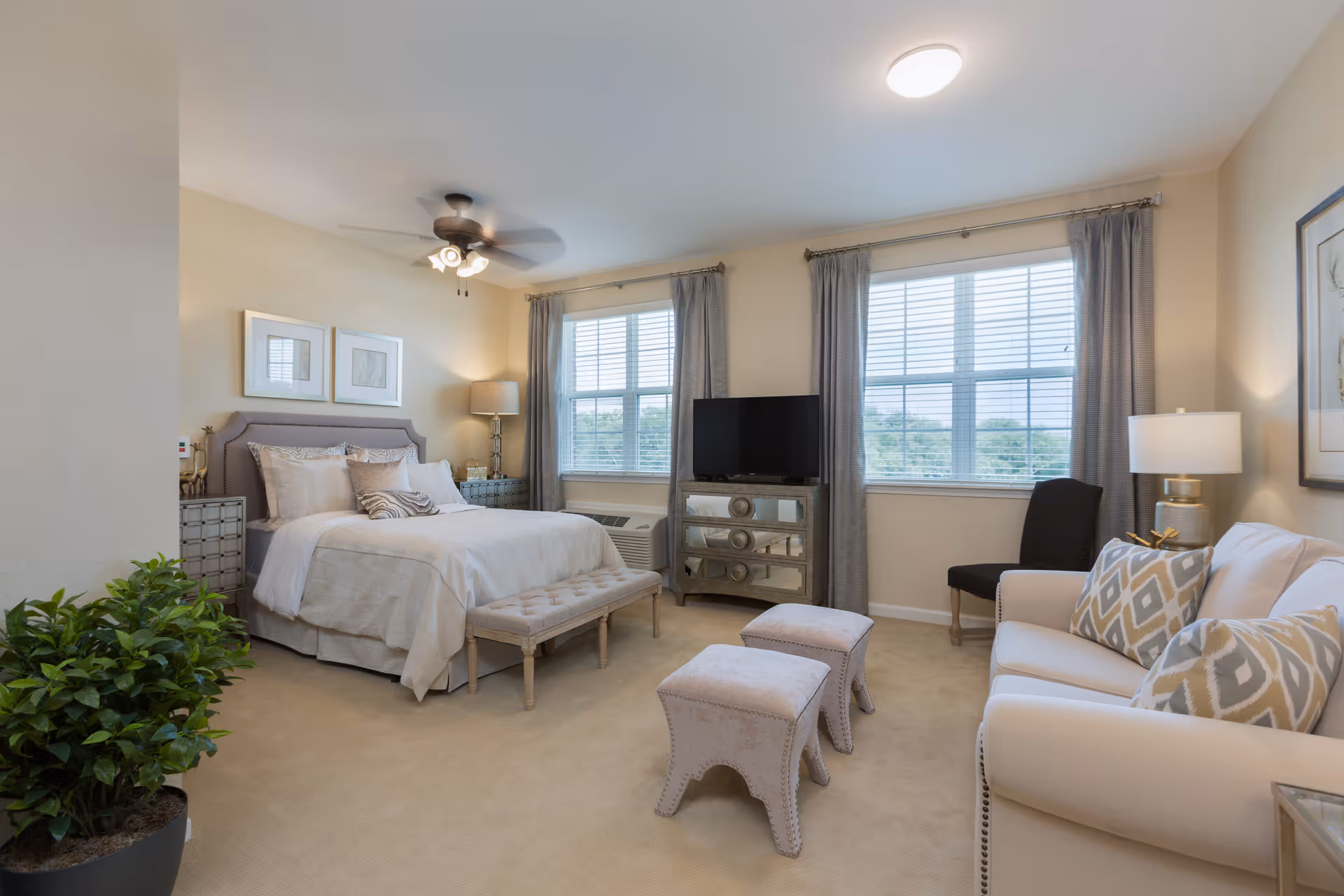 A well-lit senior living bedroom featuring a large bed with white bedding and multiple pillows, a bench at the foot of the bed, two windows with gray curtains, a TV on a mirrored dresser, a white sofa with patterned pillows, two small upholstered stools, a black chair, a potted plant, and ceiling fan with lights.
