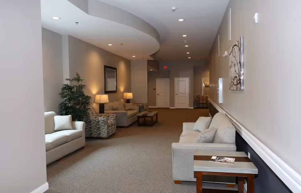 Spacious neutral-toned communal lounge with sofas, armchairs, side tables and lamps along a long hallway.
