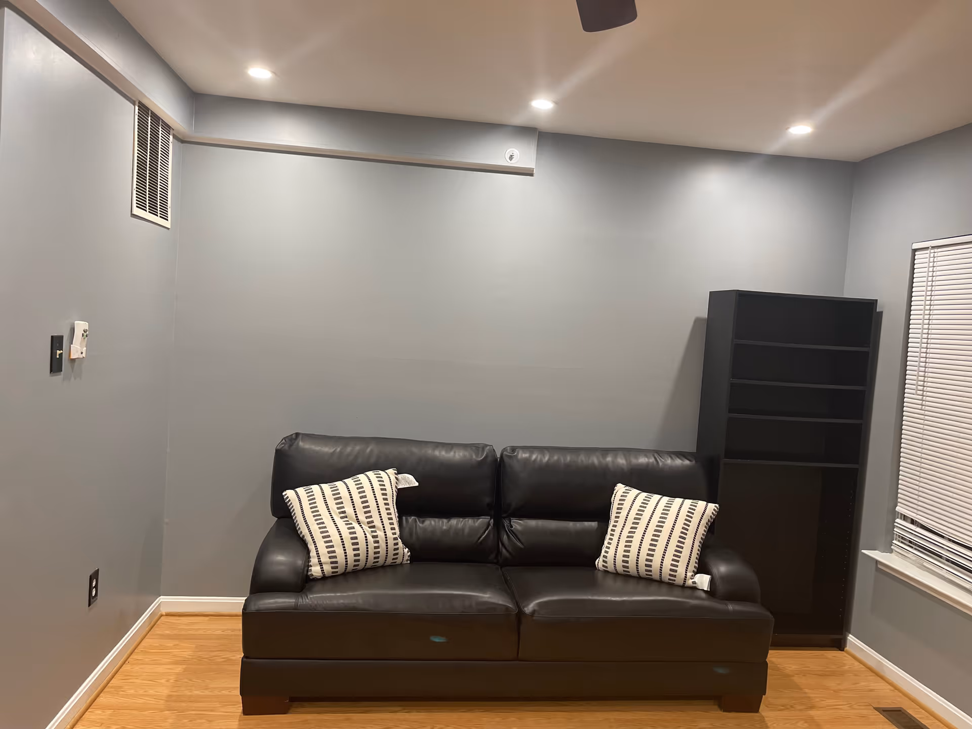 A small living room with a black leather couch adorned with two white and black patterned pillows. To the right of the couch is a tall black shelving unit, and a window with closed blinds. The walls are painted light gray and the floor is wooden. The ceiling has recessed lighting and a ceiling fan is partially visible.