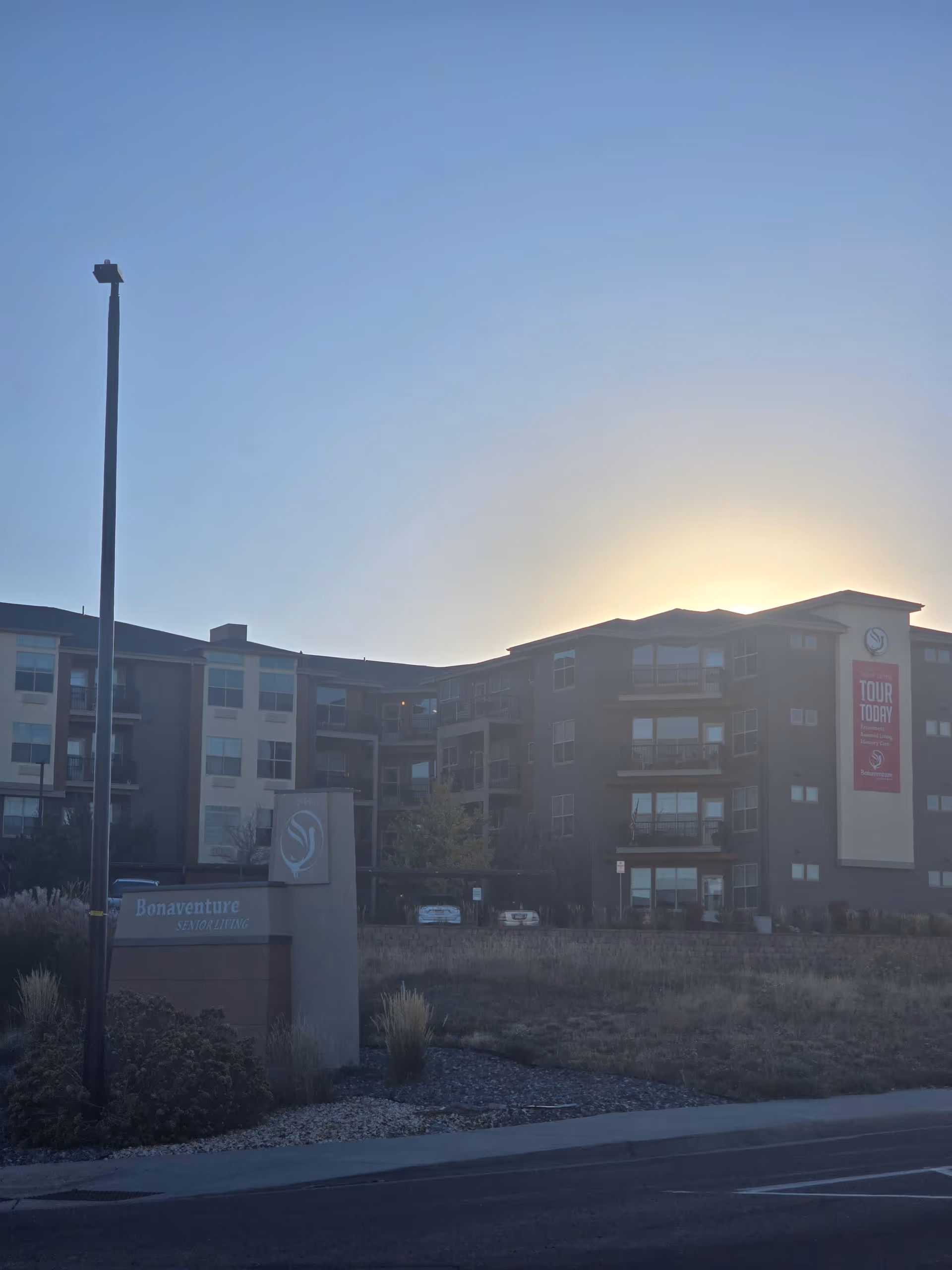 Exterior view of Bonaventure Senior Living facility at sunset with the sun setting behind the building. The multi-story building has balconies and large windows. There is a sign in front that reads 'Bonaventure Senior Living' and a tall street lamp nearby.