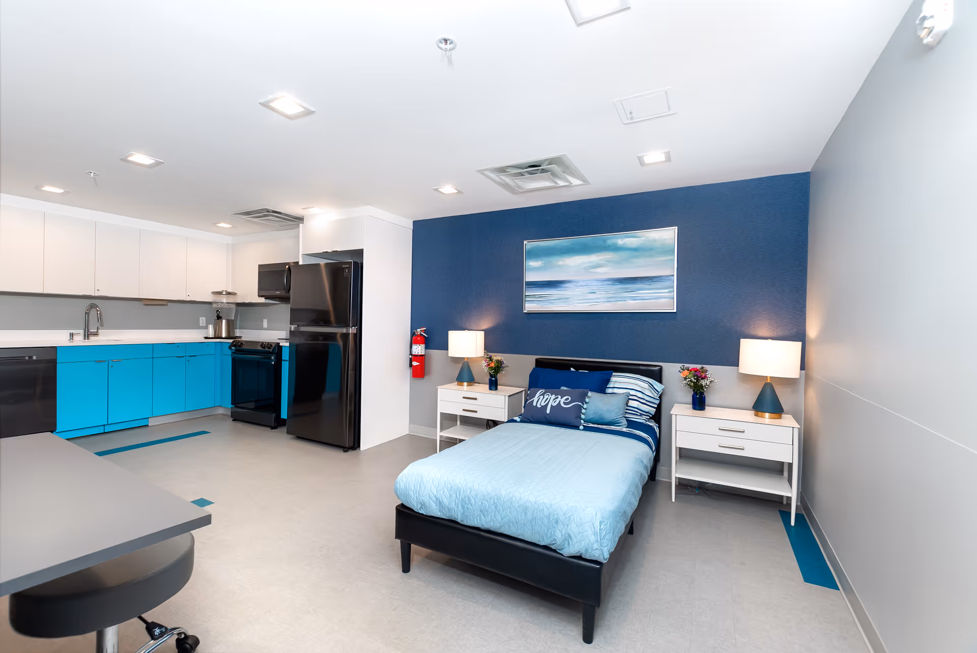A bright and modern room featuring a single bed with blue bedding and pillows, two white nightstands with lamps and flower vases, and a large painting of an ocean scene on a blue accent wall. The room also includes a kitchenette with turquoise lower cabinets, white upper cabinets, a black refrigerator, stove, and dishwasher. The floor is light-colored with blue accent tiles, and the ceiling has recessed lighting.