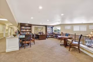 A spacious, well-lit common area with carpeted floors, several wooden tables and chairs, bookshelves filled with books, and large windows with blinds. The room has a cozy and inviting atmosphere, suitable for reading or socializing.