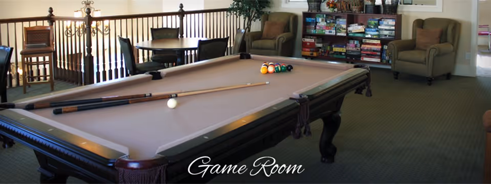 A game room featuring a pool table with cues and balls, seating, a small table, and shelves of board games.