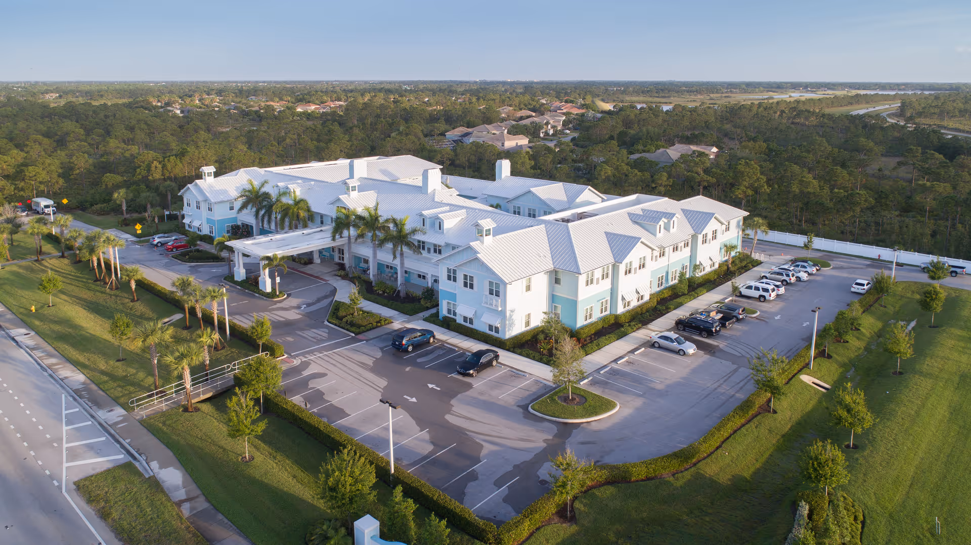 Aerial view of a two-story coastal-style senior living building with parking lot, palm trees, and landscaped grounds.