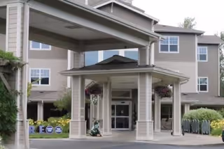 Entrance of a multi-story senior living facility with a covered drop-off area supported by columns. The building has beige siding and multiple windows. There are hanging flower baskets and greenery around the entrance.