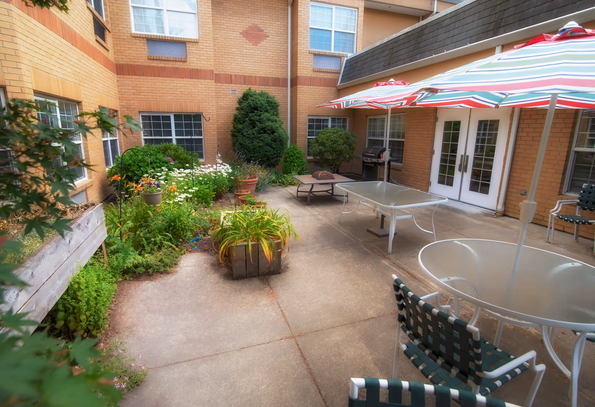 Brick-walled courtyard patio with glass tables, striped umbrellas, lawn chairs, and potted plants.