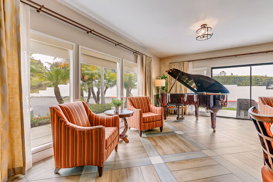 A bright and spacious living room area with large windows showing palm trees outside. The room features two striped orange armchairs with a small round wooden table between them, a grand piano, a floor lamp, and sliding glass doors leading to an outdoor patio.