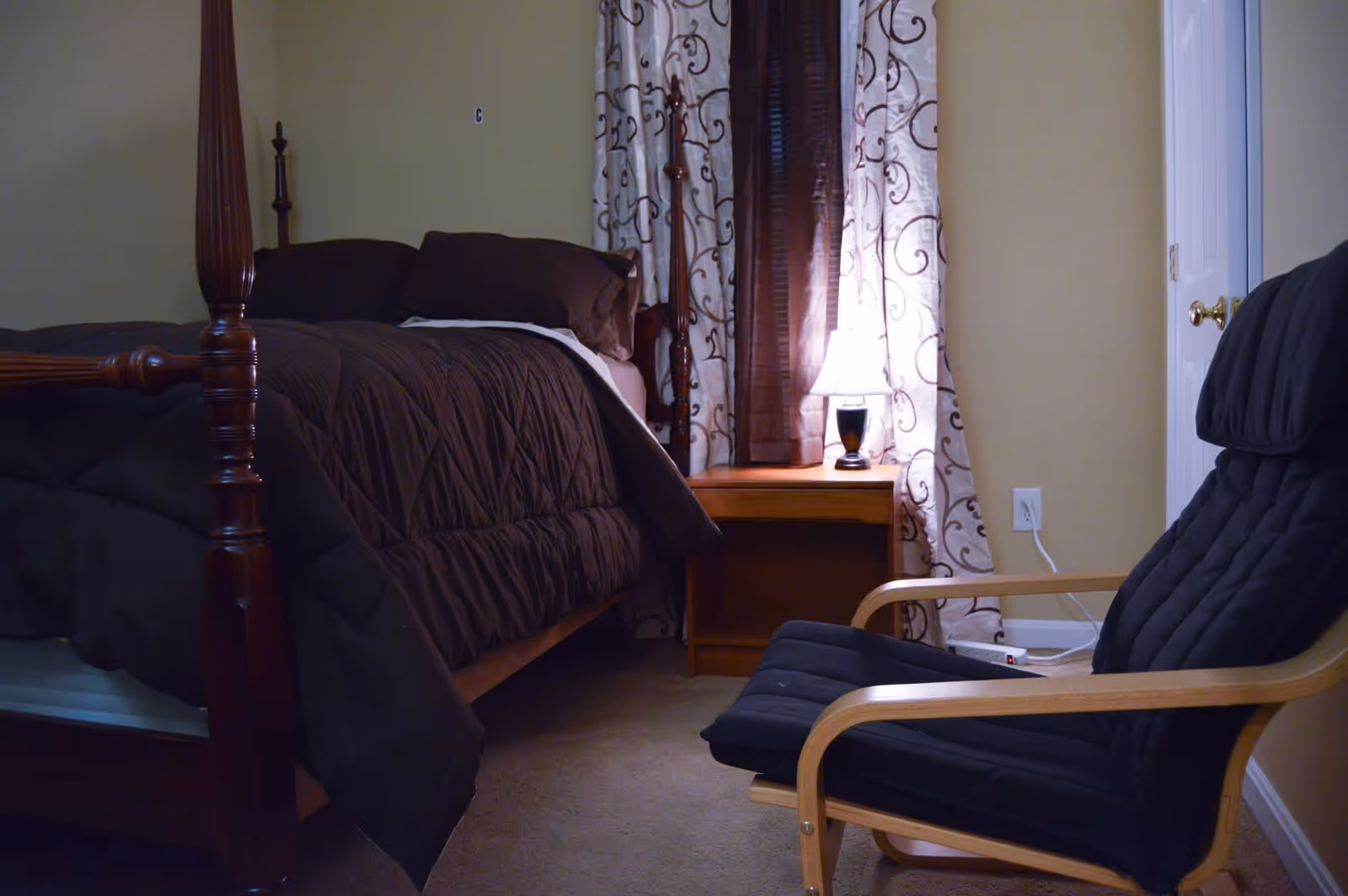 Cozy bedroom with a dark wood four‑poster bed, nightstand with a lit lamp and patterned curtains, and a black cushioned armchair.