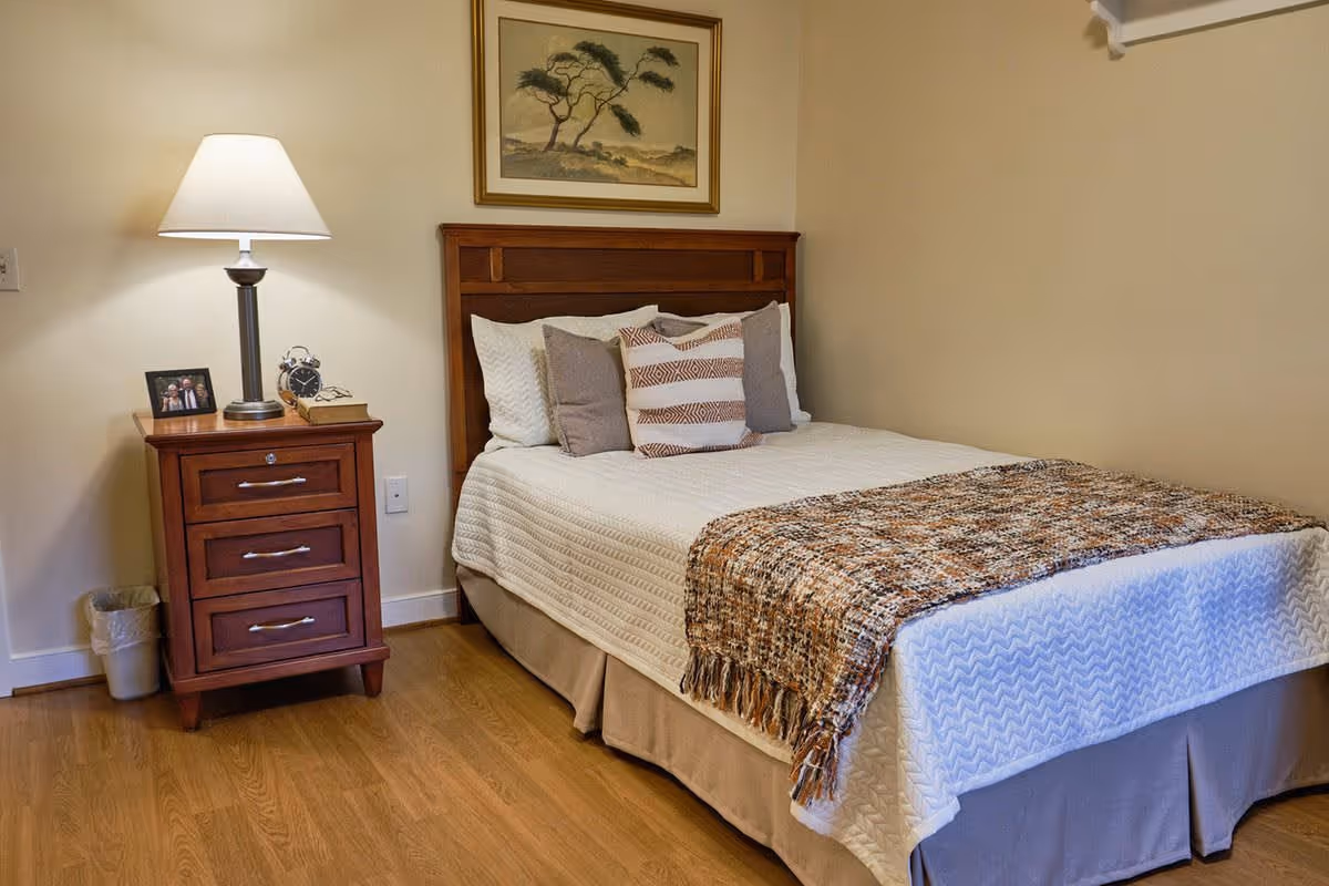 A cozy bedroom with a wooden bed frame, a neatly made bed with white and beige bedding, and several decorative pillows. Next to the bed is a wooden nightstand with three drawers, a table lamp, a framed photo, an alarm clock, and a book. A framed painting of a tree hangs on the wall above the bed. The room has light-colored walls and wooden flooring.
