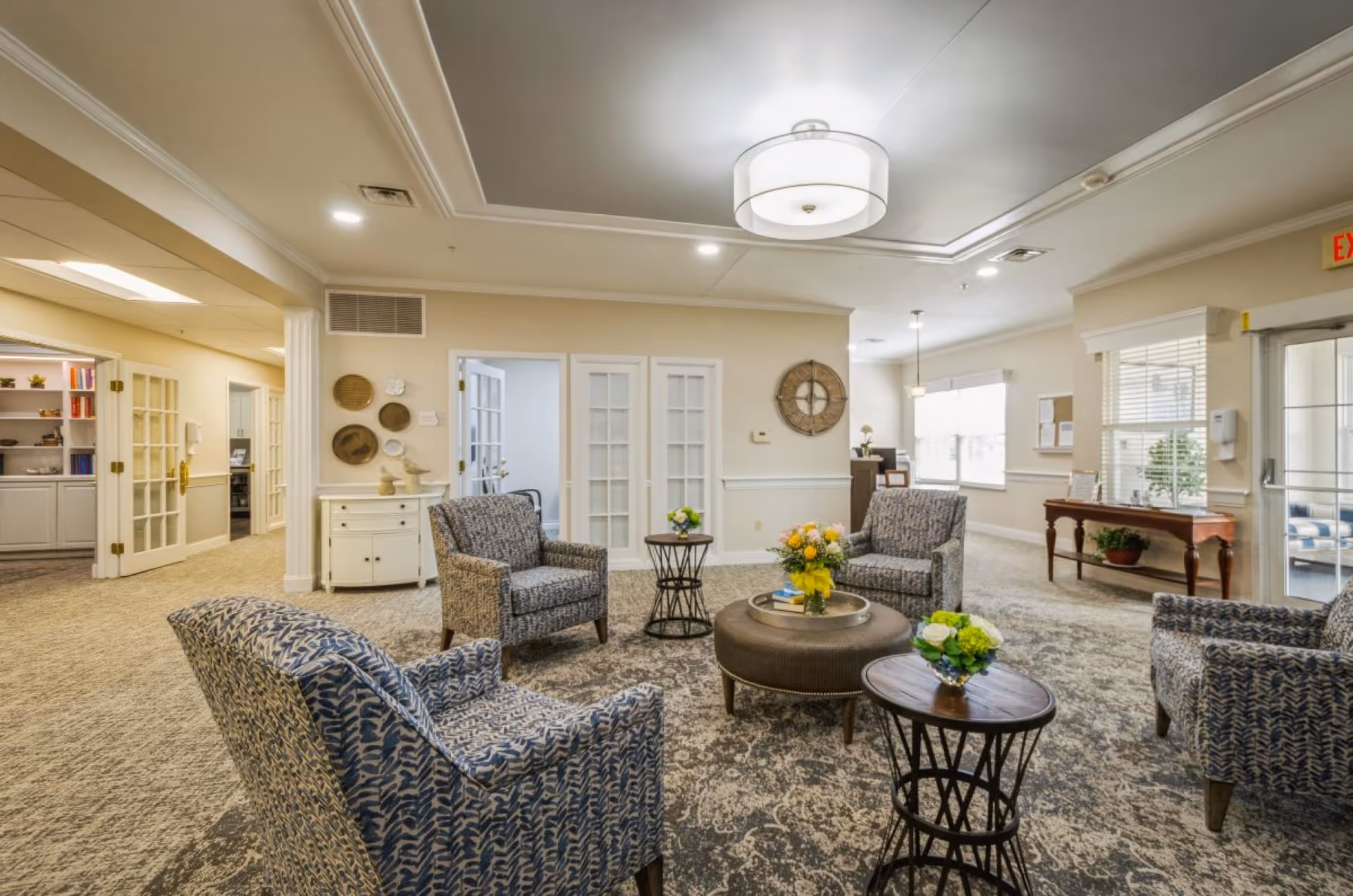 A bright and spacious sitting area in a senior living facility with four patterned armchairs arranged around a round ottoman and two small side tables, each decorated with flower arrangements. The room has beige walls, carpeted floors, a modern ceiling light fixture, and French doors leading to other rooms. There is a wooden console table with plants and decor near a window, and an exit door is visible on the right side.