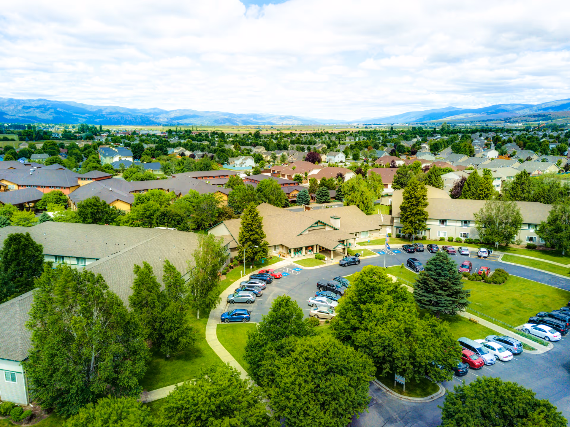 Aerial view of Missoula Valley Senior & Assisted Living facility showing multiple buildings surrounded by green trees and lawns, with a parking lot filled with cars. The background includes a suburban neighborhood and distant mountains under a partly cloudy sky.