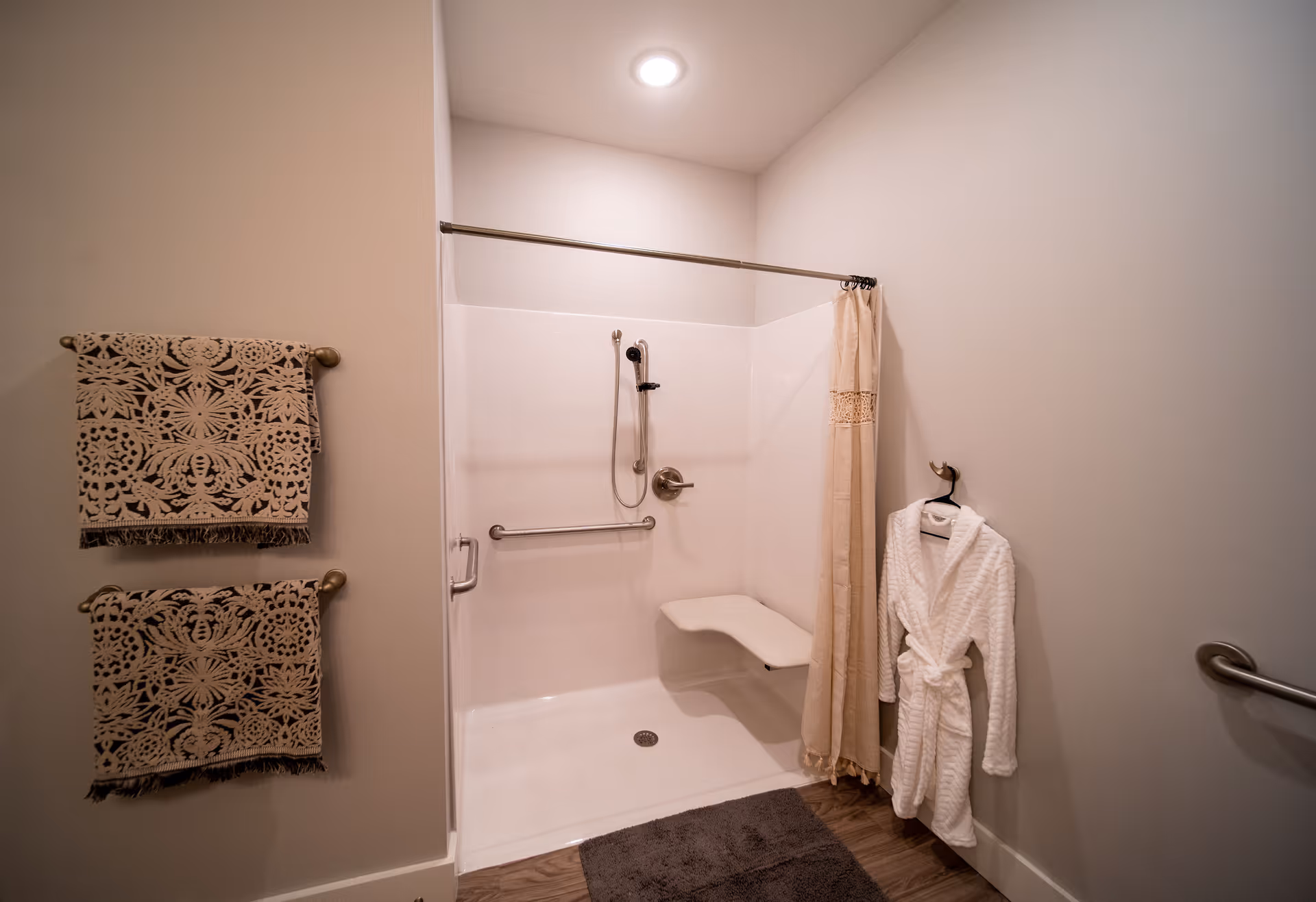 A clean, accessible shower area with a fold-down seat and grab bars. The shower has a handheld showerhead and a beige curtain partially drawn to the side. Two patterned towels hang on a towel rack on the left wall, and a white bathrobe hangs on a hook on the right wall. The floor is wood-style with a dark bath mat in front of the shower.
