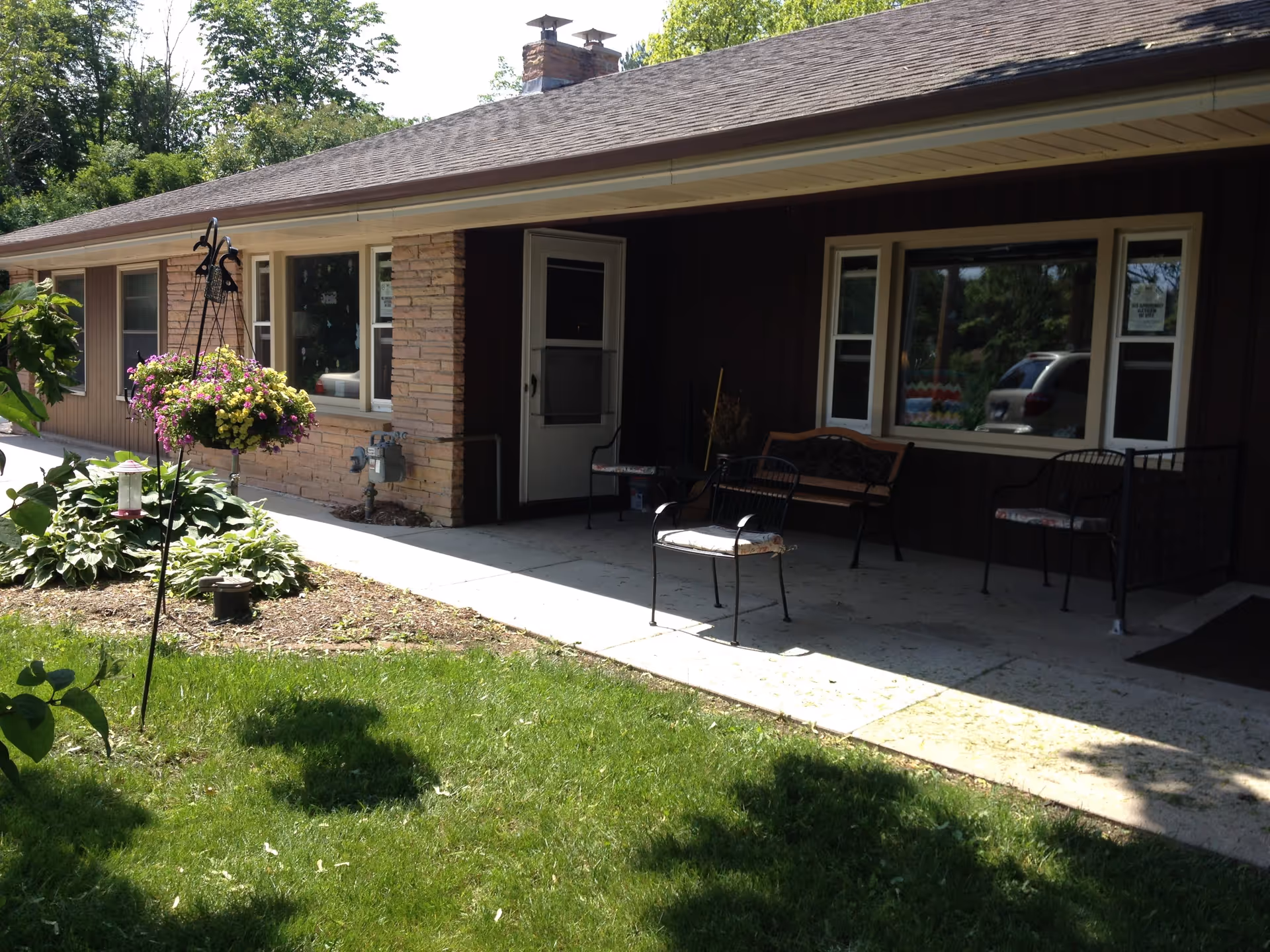 Exterior view of a single-story building with a covered porch area. The porch has several chairs and a bench, and there is a hanging flower basket with colorful flowers near the walkway. The building has large windows and a shingled roof, surrounded by green grass and trees.