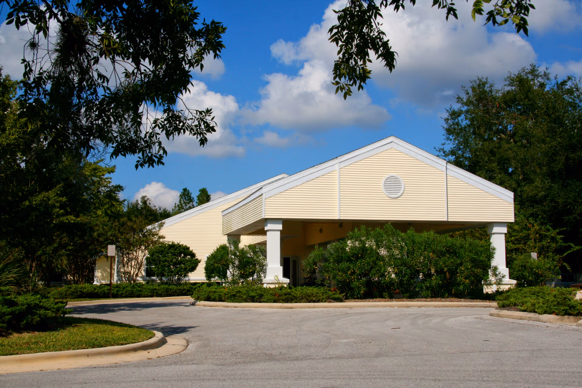 Exterior view of a single-story building with light yellow siding and a covered entrance supported by white columns. The building is surrounded by green bushes and trees under a blue sky with scattered clouds.