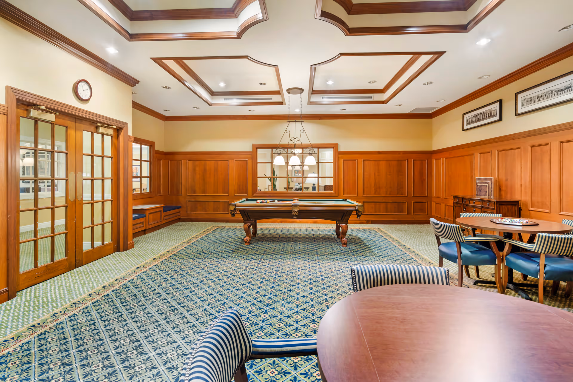 A well-lit game room with a pool table in the center, surrounded by wood-paneled walls and decorative ceiling beams. There are round tables with striped chairs on the right side, and a large patterned carpet covers the floor. Double glass doors and a clock are visible on the left wall.