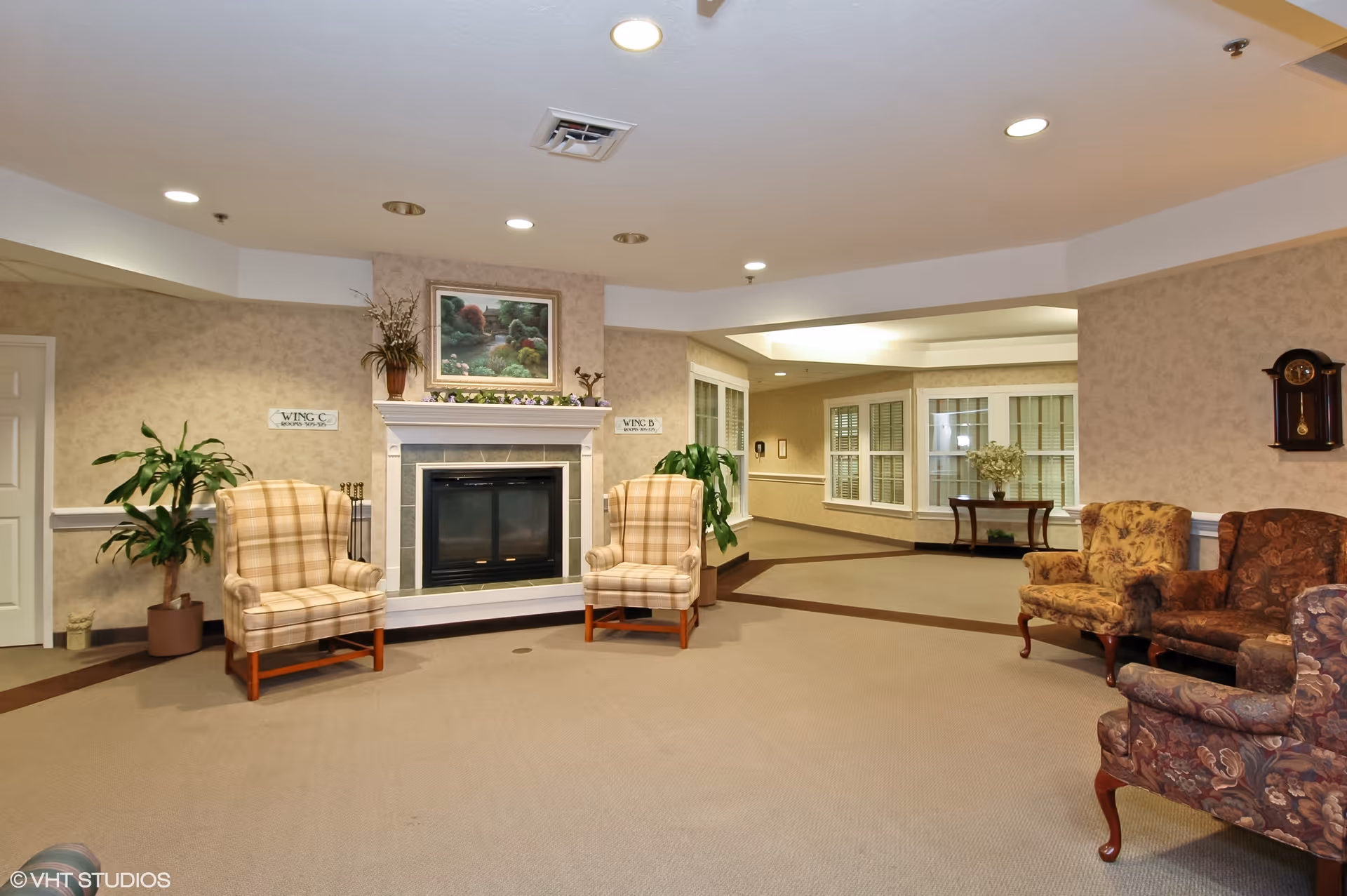A cozy common area in a senior living facility featuring a fireplace with a painting above it, two plaid armchairs on either side of the fireplace, and several floral-patterned armchairs arranged around the room. There are potted plants, a wall clock, and a table with a vase of flowers in the background. The walls are beige with a subtle pattern, and the ceiling has recessed lighting.