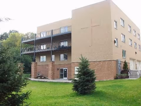 Three-story tan rehabilitation building with exterior balconies, a large cross on the facade, and a grassy lawn with small evergreen trees in front.
