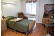 A single hospital-style bed with green bedding in a well-lit room. Next to the bed is a brown recliner chair and a wooden bedside table with a small plant and a box of tissues. The room has a wooden floor, a window with floral curtains, a wooden dresser with plants and picture frames, and a small round table with chairs.