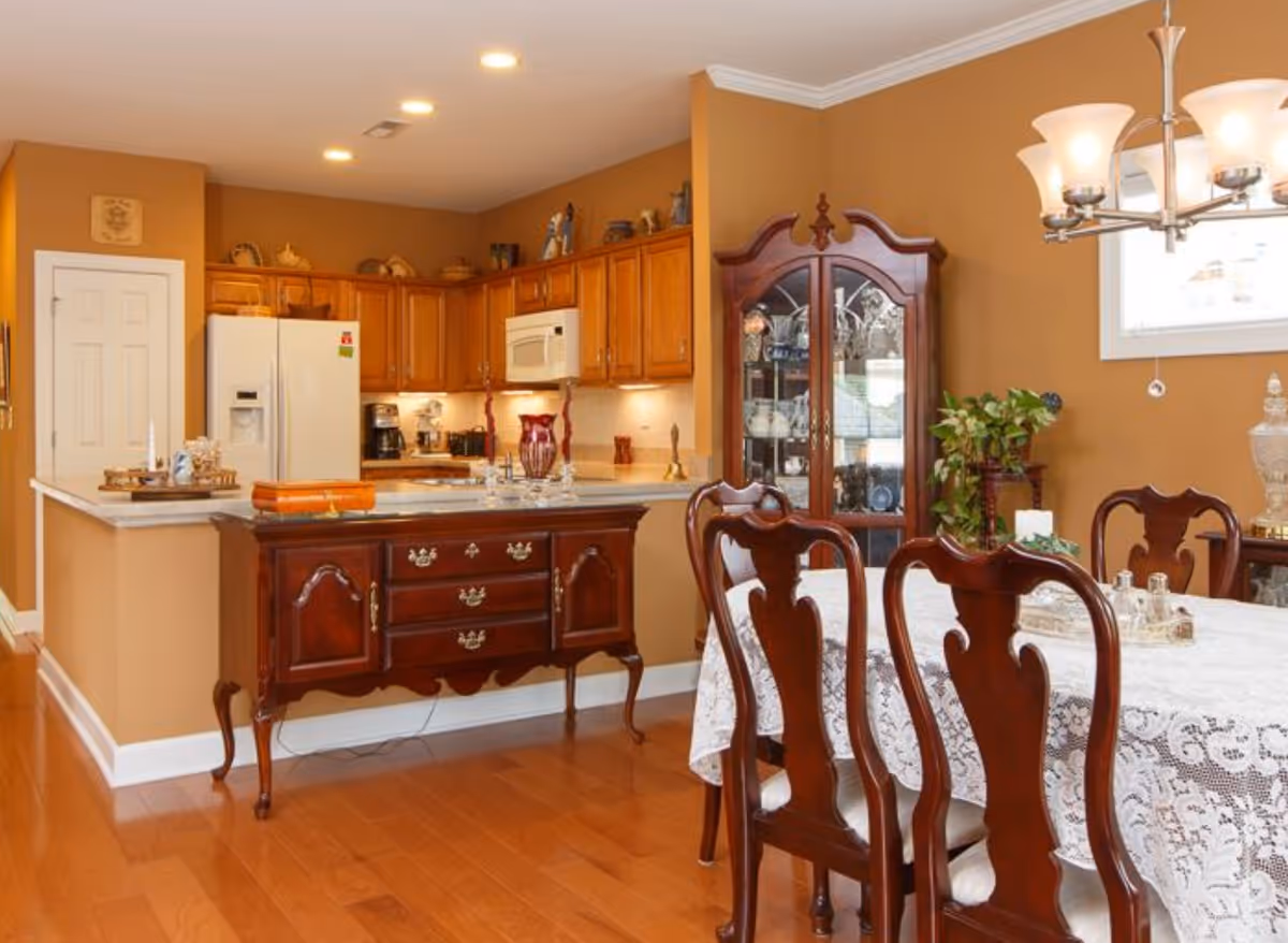 A warm-toned dining area and kitchen with wooden floors and cabinetry. The dining area features a table covered with a white lace tablecloth and dark wooden chairs. A glass-front china cabinet and a chandelier are visible. The kitchen has a white refrigerator, microwave, and various kitchen appliances on the countertops. A wooden sideboard with decorative items is positioned between the dining area and kitchen.