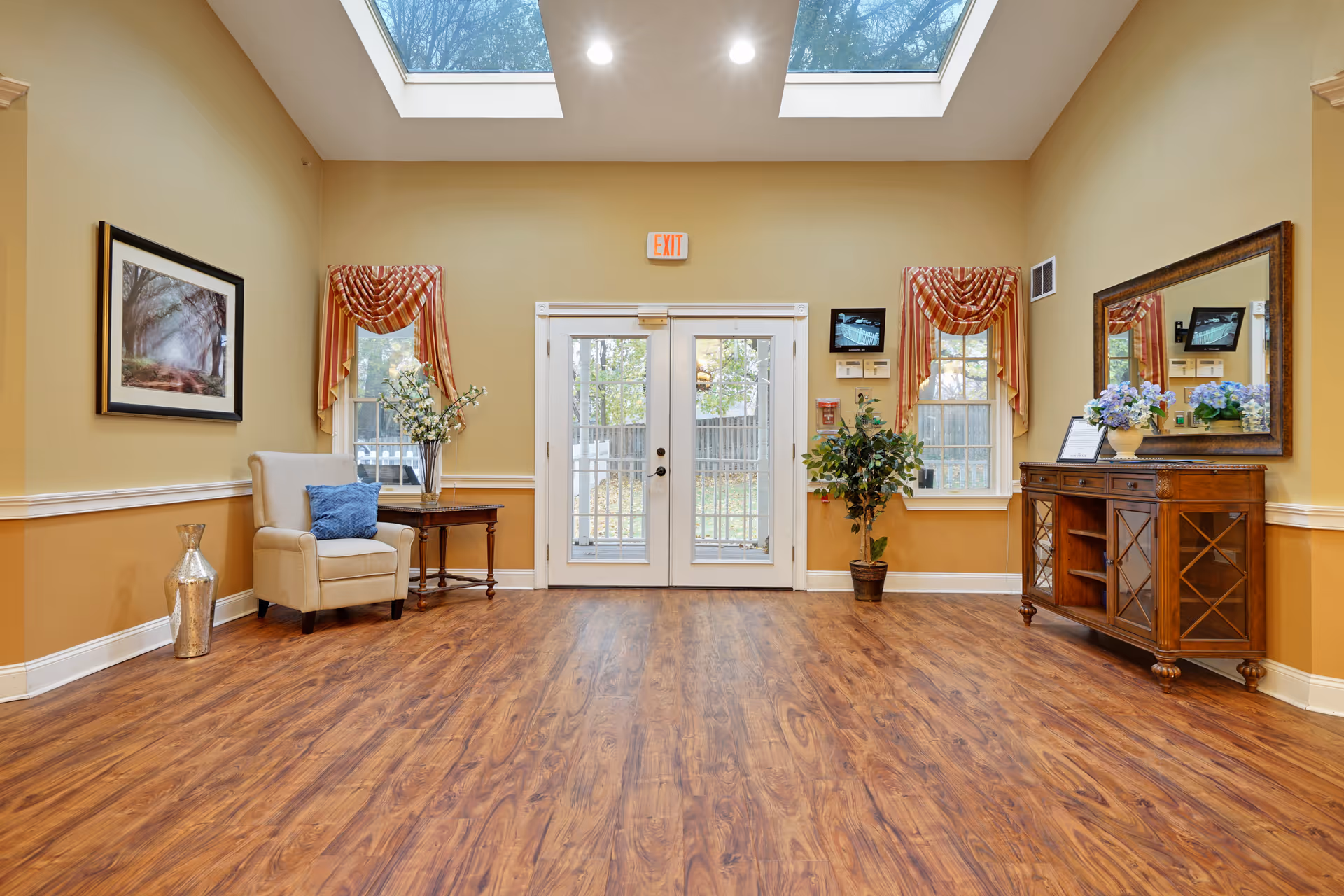 A bright and spacious interior room with wooden flooring, beige walls, and a high ceiling with skylights. The room features a beige armchair with a blue cushion next to a small wooden table with a vase of flowers. There are two windows with red and gold drapes flanking a set of double glass doors leading outside. On the right side, there is a wooden sideboard with a large mirror above it and a vase of flowers on top. A potted plant is placed near the right window. The room is well-lit with natural light from the skylights and windows.