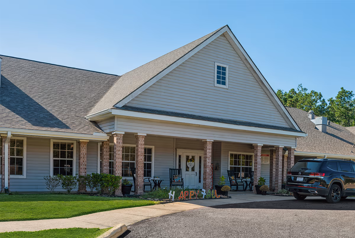 Exterior view of a single-story building with a covered porch supported by brick columns. The porch has chairs and small tables, and a decorative sign on the ground reads 'Happy Fall'. A black SUV is parked near the entrance. The building is surrounded by green grass and trees under a clear blue sky.