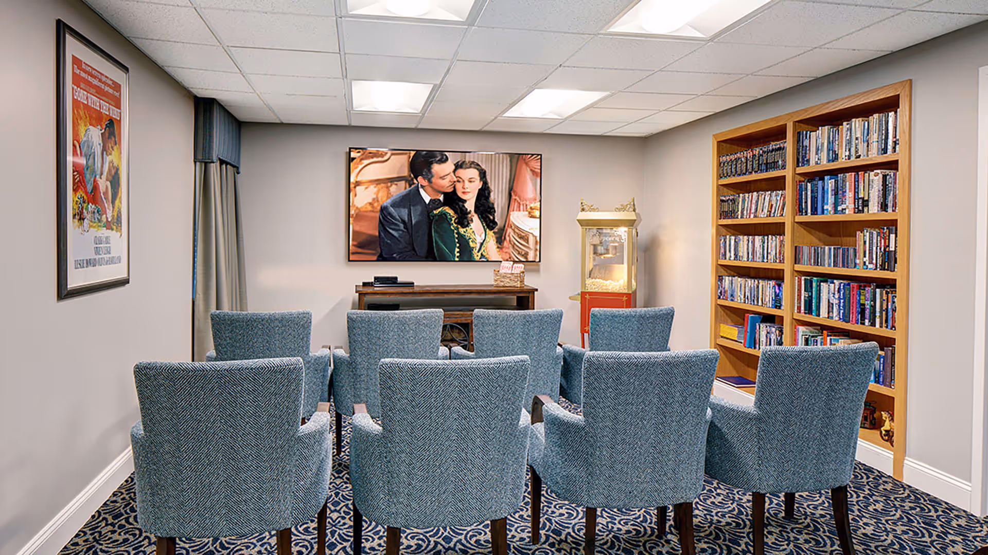 A small movie screening room with eight blue upholstered chairs arranged in two rows facing a wall-mounted TV showing a classic movie scene. To the right, there is a wooden bookshelf filled with DVDs and books, and a popcorn machine is placed next to the TV. The room has patterned carpet and light-colored walls with a framed movie poster on the left wall.