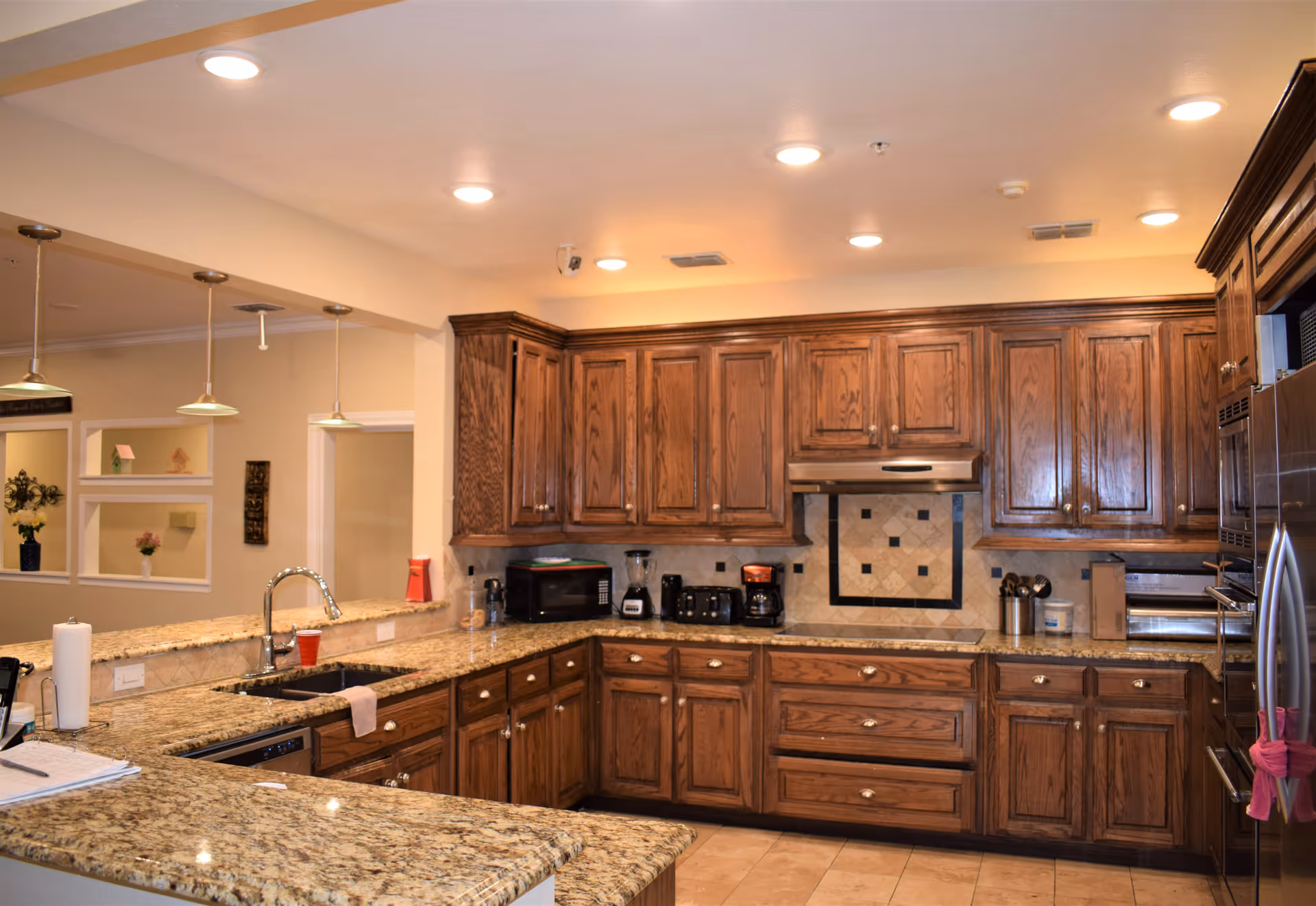 A spacious kitchen with wooden cabinets and granite countertops. The kitchen features a double sink, a microwave, a blender, a coffee maker, a toaster, and a stainless steel refrigerator. There are pendant lights hanging over the counter and recessed lighting in the ceiling. The backsplash has a decorative tile design behind the stove area.