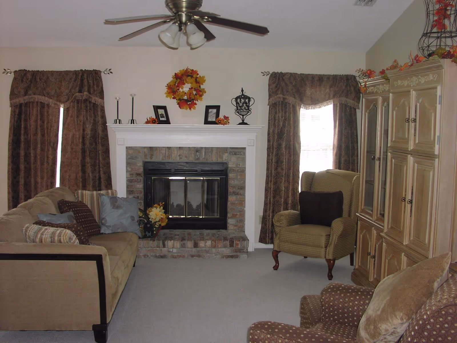 A cozy living room with a beige sofa adorned with multiple pillows, a patterned armchair, and a wooden cabinet. The room features a brick fireplace with a white mantel decorated with a fall-themed wreath, candles, and framed pictures. Two windows with brown patterned curtains flank the fireplace, and a ceiling fan with lights is mounted on the ceiling.