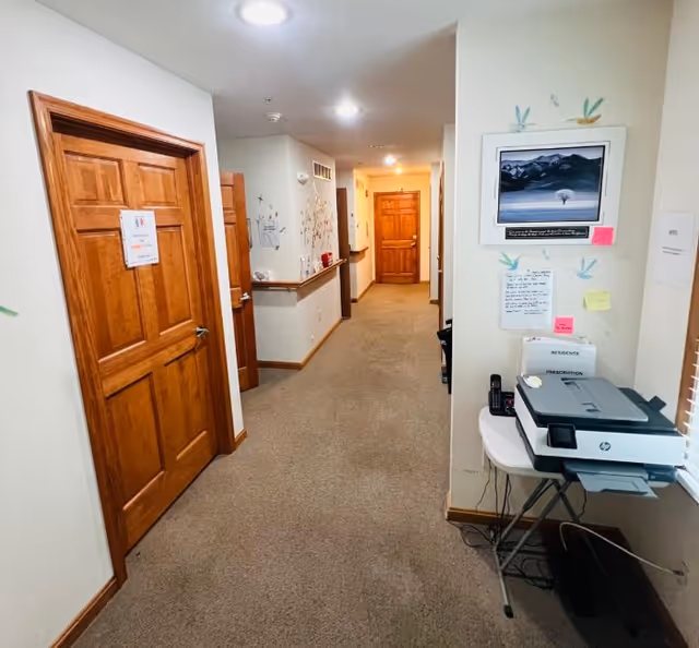 Carpeted interior hallway in an assisted living facility with wooden doors, wall art, and a small printer station.