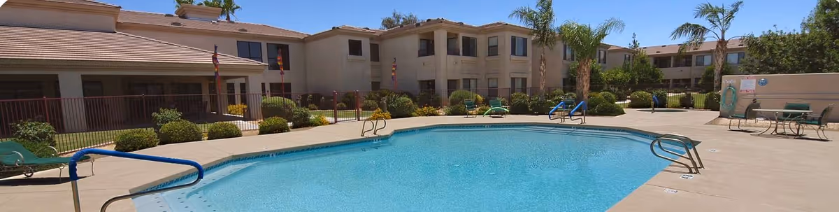 Outdoor swimming pool surrounded by lounge chairs and patio furniture in front of a two-story beige senior living building.