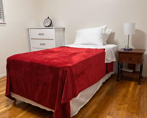 A simple bedroom with a single bed covered with a red blanket and white pillows. Next to the bed is a wooden nightstand with a white lamp and a small clock. A white dresser with a round clock on top is against the wall. The room has wooden flooring and white walls.