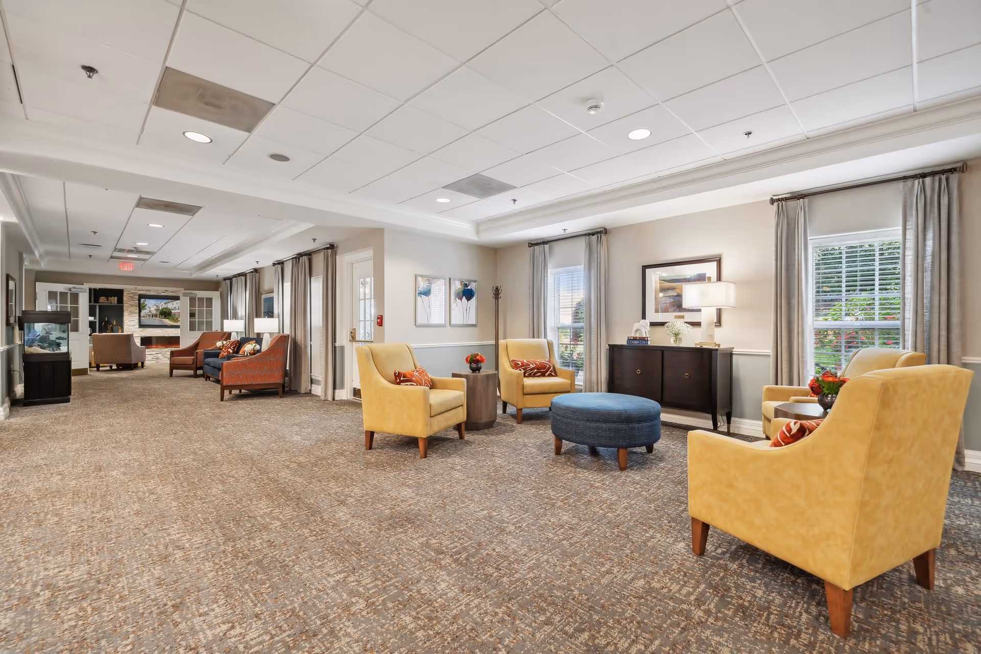 Bright, spacious common seating area with yellow armchairs, a blue ottoman, side tables, and a TV/lounge area in the background.