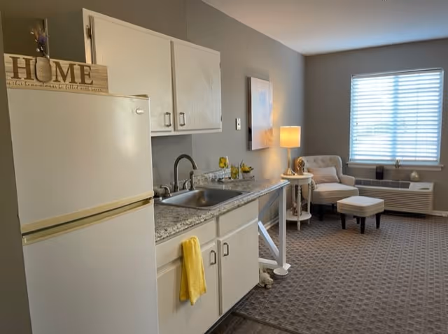 A small kitchen area with a white refrigerator, white cabinets, and a granite countertop with a sink. A yellow towel hangs from the cabinet handle. To the right, there is a cozy living area with a beige armchair, matching ottoman, a side table with a lamp, and a window with blinds letting in natural light.