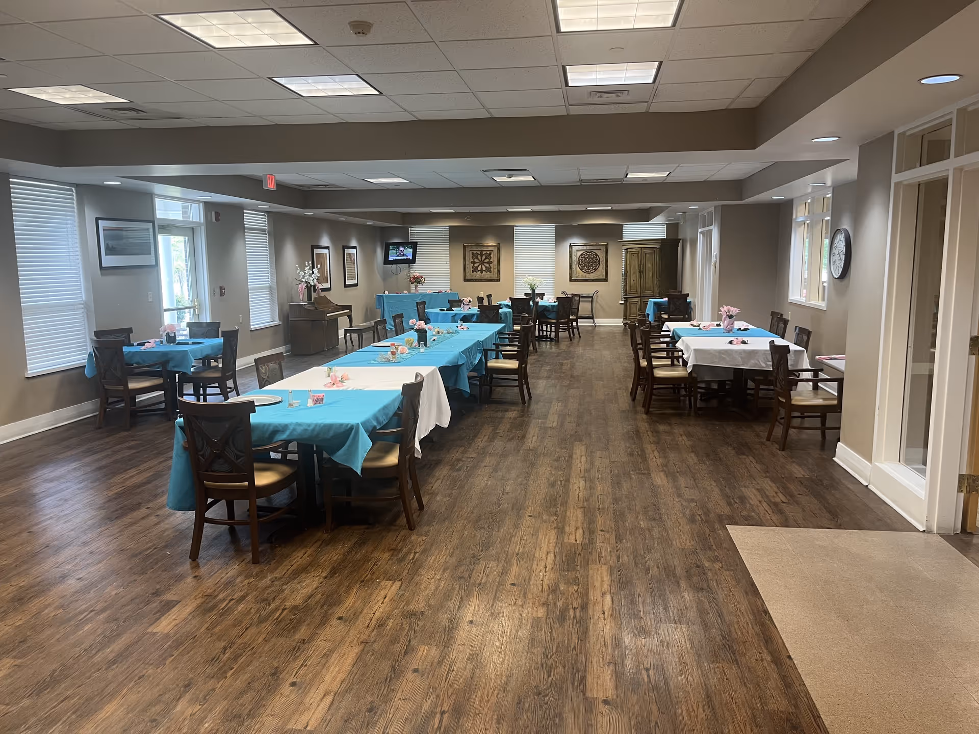 A spacious dining room with several tables covered in blue and white tablecloths, each set with chairs and small floral centerpieces. The room has wooden flooring, beige walls, multiple windows with blinds, and ceiling lights. There is a piano against one wall and a wall-mounted TV in the background.