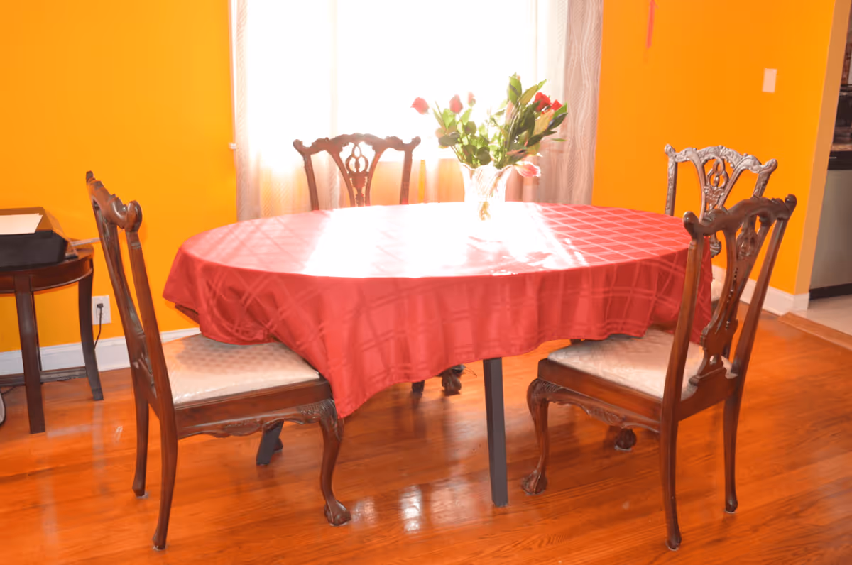 Oval dining table covered with a red tablecloth surrounded by four ornate wooden chairs in a bright orange-walled room with a vase of flowers.