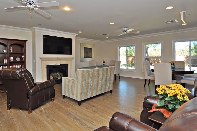 A spacious living room area with wooden flooring, featuring a striped sofa, two leather armchairs, a fireplace with a mounted flat-screen TV above it, a wooden bookshelf, and a dining table with upholstered chairs near large windows letting in natural light.