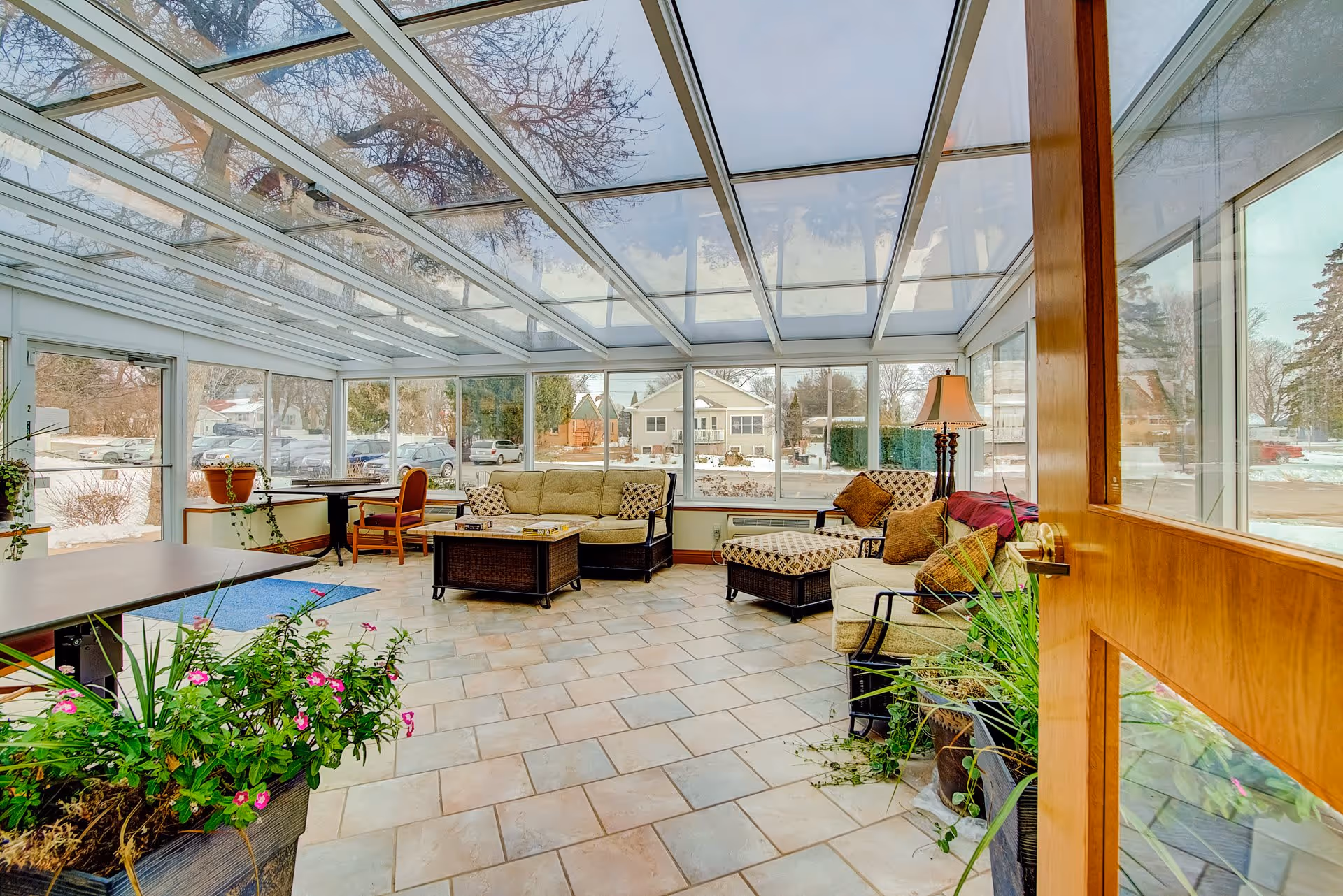 Bright glass-enclosed sunroom lounge with tiled floor, sofas, chairs, plants and a view of the snowy exterior.