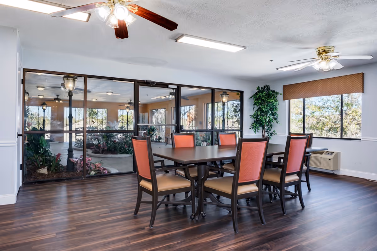 A dining area with a rectangular table and six chairs on dark wood flooring, ceiling fans, large windows, and an indoor garden visible through glass panels.