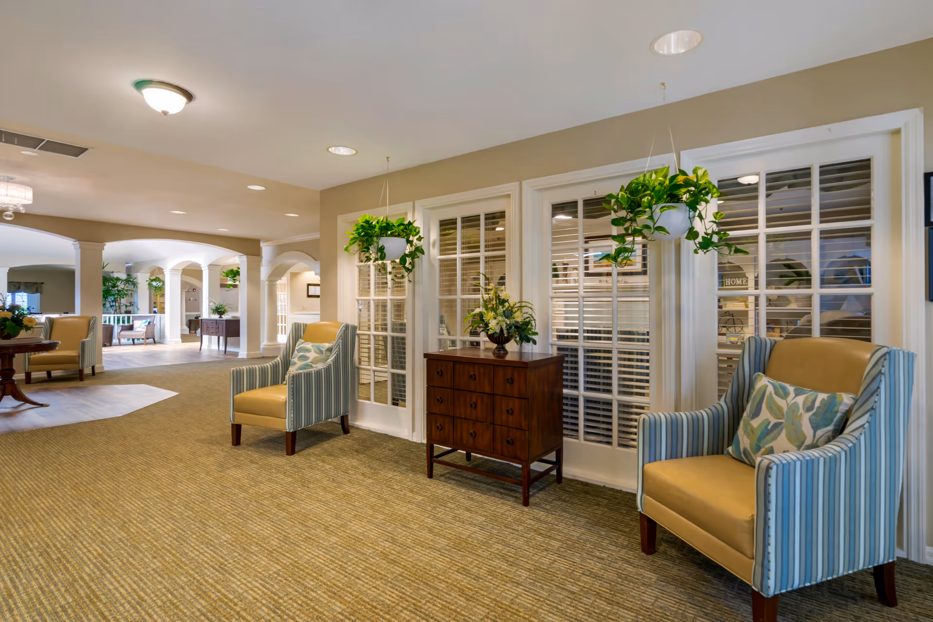 A bright senior living lounge with two striped armchairs, hanging plants, a wooden chest, and large interior windows opening to a columned common area.