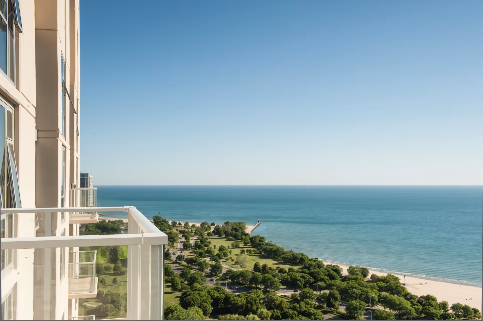 View from a high balcony of a building overlooking a green park area with trees and a beach along a large body of water under a clear blue sky.