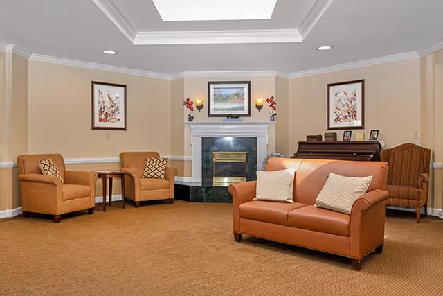 A cozy living room area with a brown leather sofa, two matching armchairs, a fireplace with a framed picture above it, and a piano in the corner. The walls are beige with white trim, and there are two framed artworks on either side of the fireplace. The room has recessed lighting and a skylight.