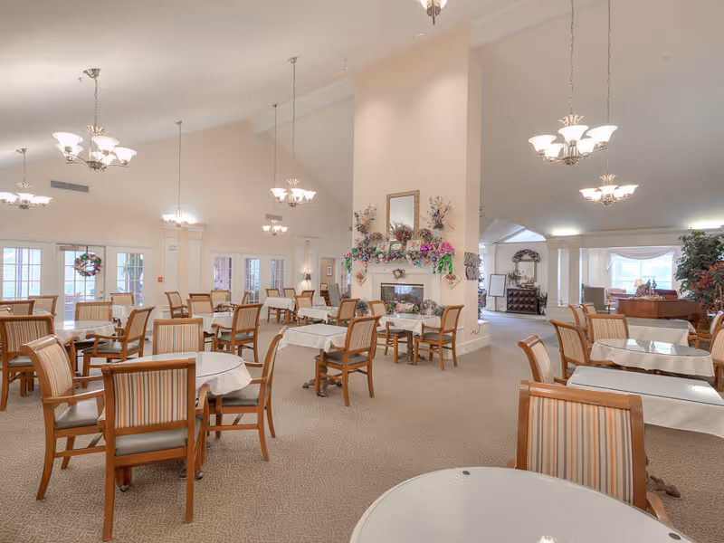 Spacious dining room with multiple round and rectangular tables and chairs, chandeliers overhead, and a decorated fireplace.