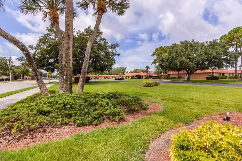 A landscaped outdoor area with green grass, shrubs, and several palm trees. In the background, there are single-story buildings with red roofs and large trees under a partly cloudy sky.