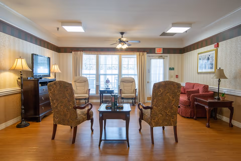 A cozy living room area in a senior living facility with two patterned armchairs facing a wooden coffee table, two beige cushioned chairs near a large window with curtains, a red sofa with matching pillows, a wooden side table with a lamp and plant, a wooden dresser with a flat-screen TV and two lamps, and a ceiling fan with lights. The room has wood flooring and wallpapered walls with a decorative border.