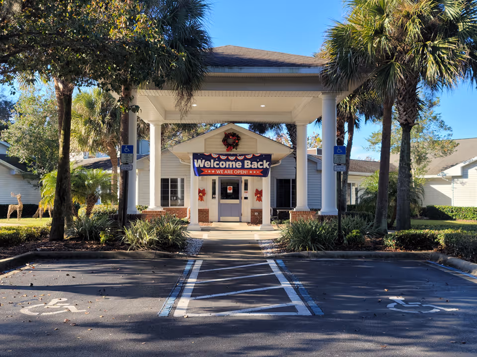 Entrance of a senior living facility with a covered porch supported by white columns, a 'Welcome Back WE ARE OPEN!' banner hanging above the door, surrounded by palm trees and landscaping, with two handicap parking spaces in front.