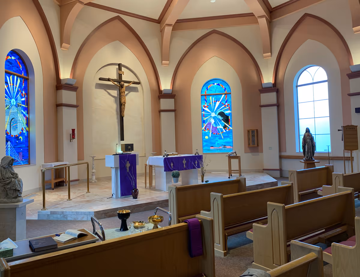 Interior of a chapel with wooden pews, a crucifix mounted on the wall behind the altar, stained glass windows, religious statues, and purple cloths draped over the altar and lectern.