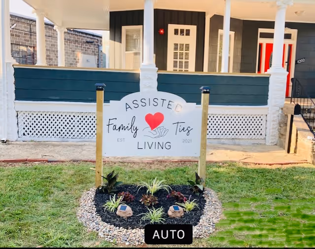 Front exterior view of a building with a porch and a sign in the lawn that reads 'Assisted Family Ties Living EST 2021' with a red heart and hands graphic.