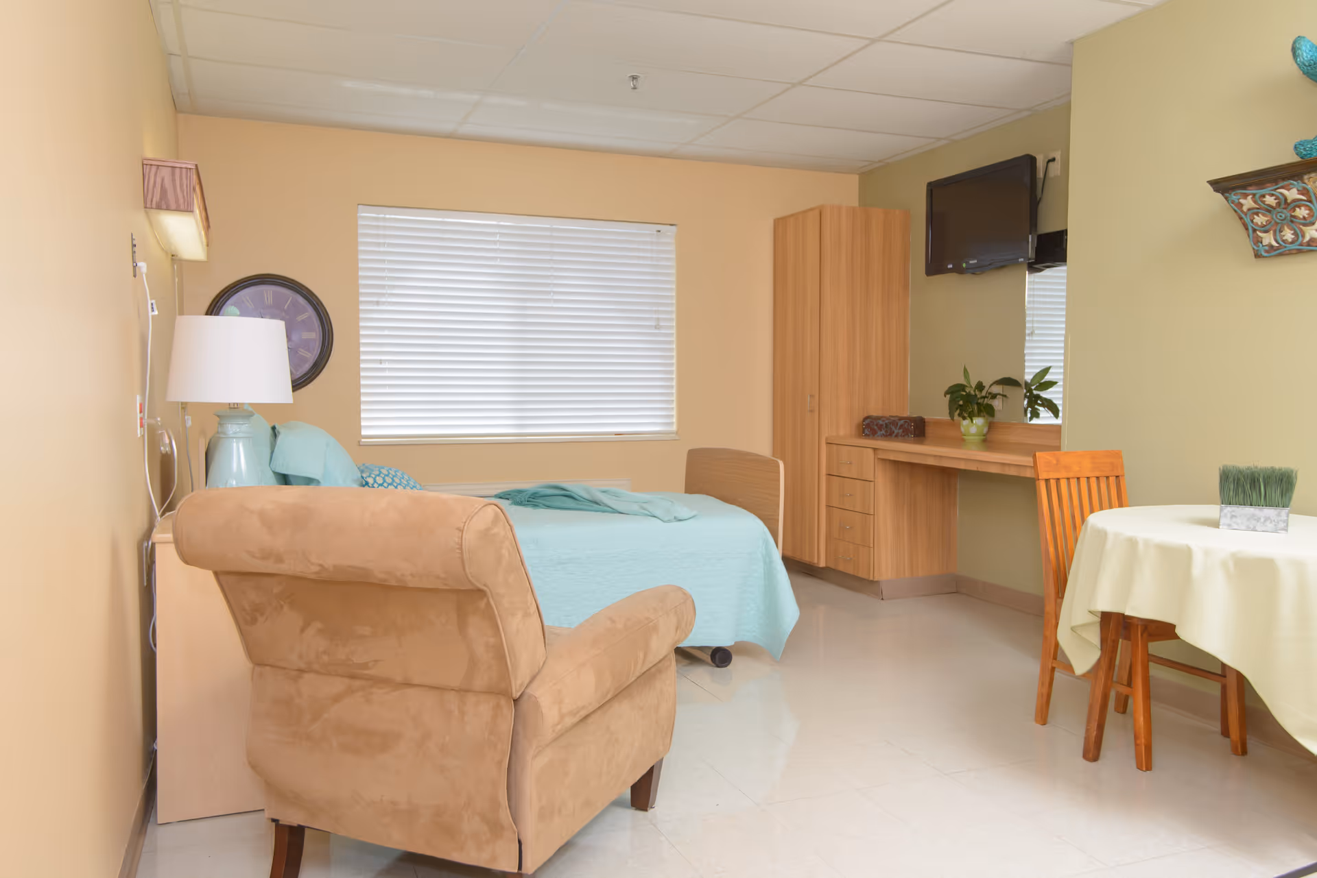 Furnished senior living bedroom with a bed, armchair, desk with a wall-mounted TV, and a small dining table.