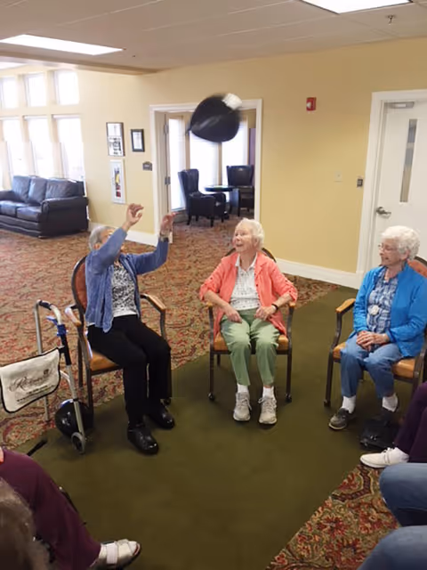 A group of elderly women sitting in a circle in a common area of a senior living facility. One woman is tossing a black hat into the air while the others watch and smile. The room has beige walls, patterned carpet, and large windows letting in natural light. There are chairs and a walker visible nearby.